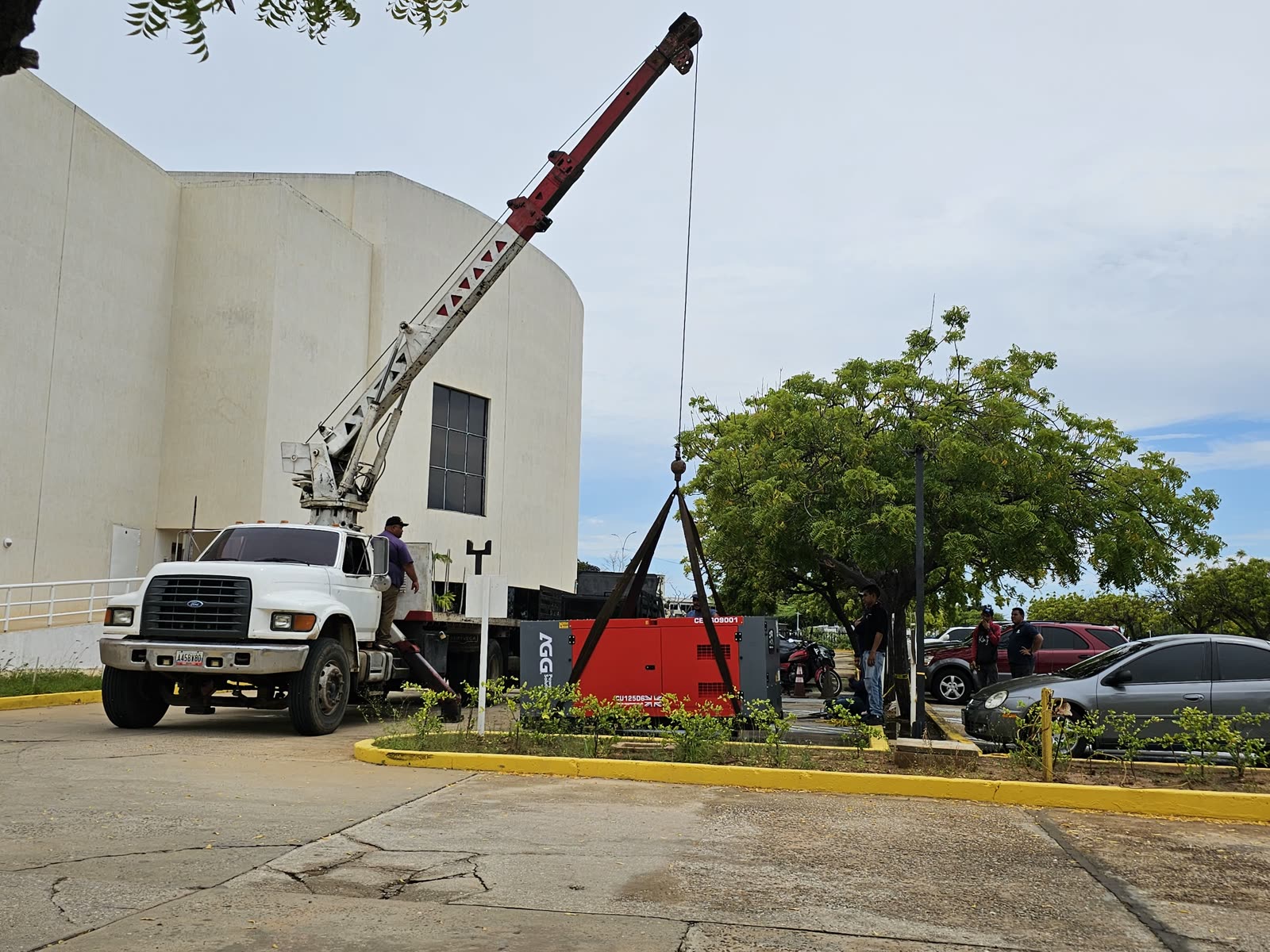 Camión grúa colocando un generador industrial rojo junto a edificio institucional